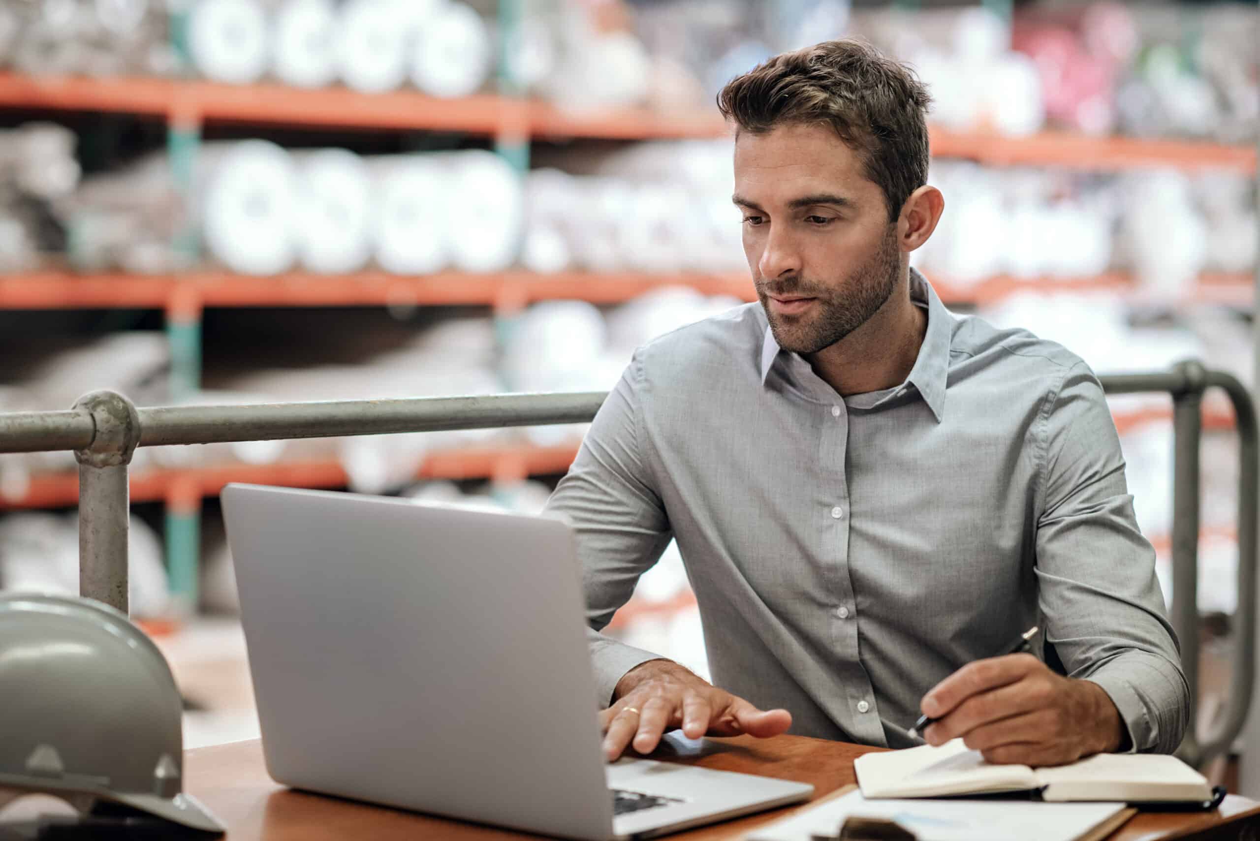 Warehouse manager taking notes and checking orders with a laptop Manager checking online orders and inventory on a laptop while sitting at a desk in a carpet warehouse with shelves of stock in the background.