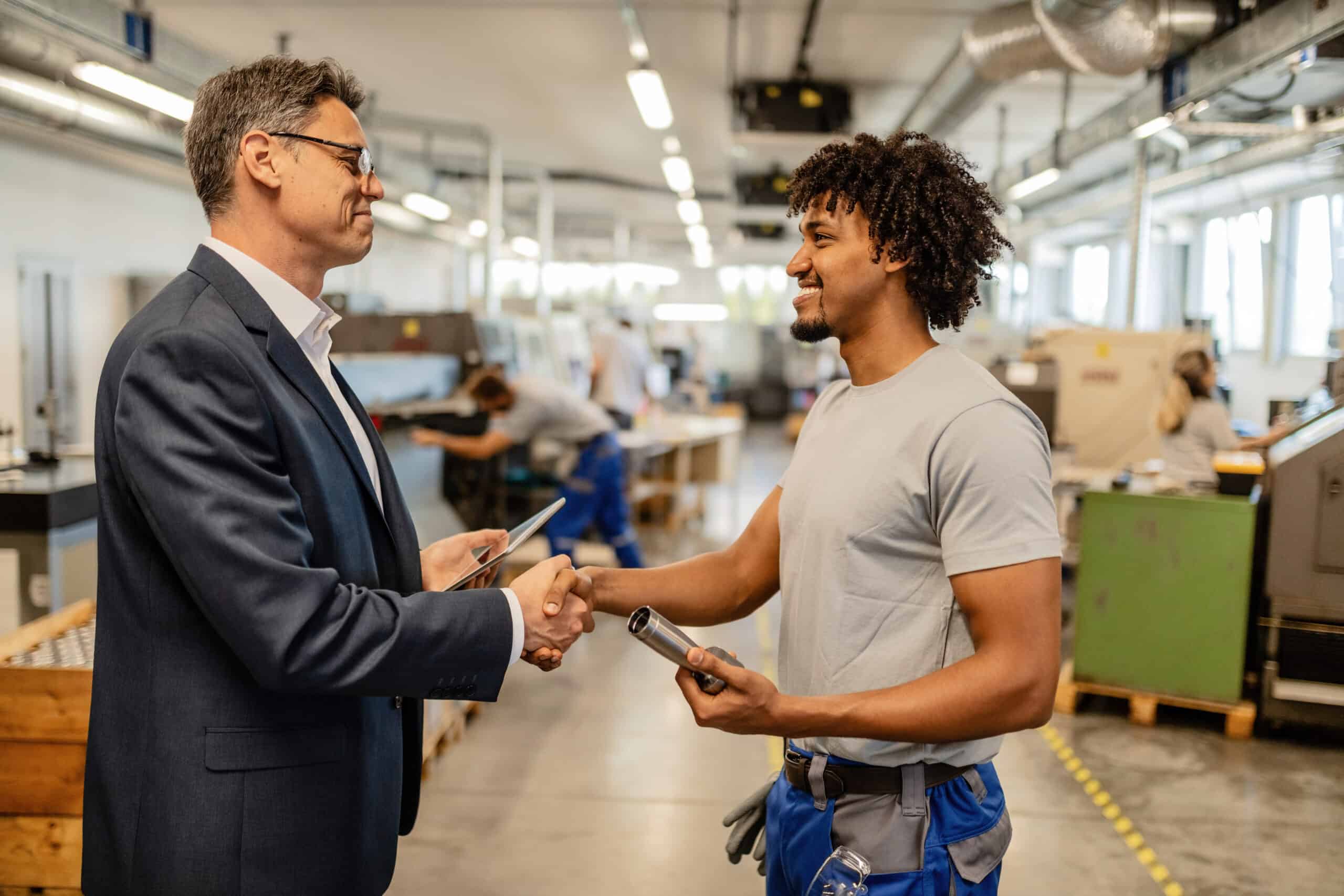 Happy businessman and African American manual worker shaking hands in factory plant. Happy businessman and African American manual worker shaking hands in factory plant.