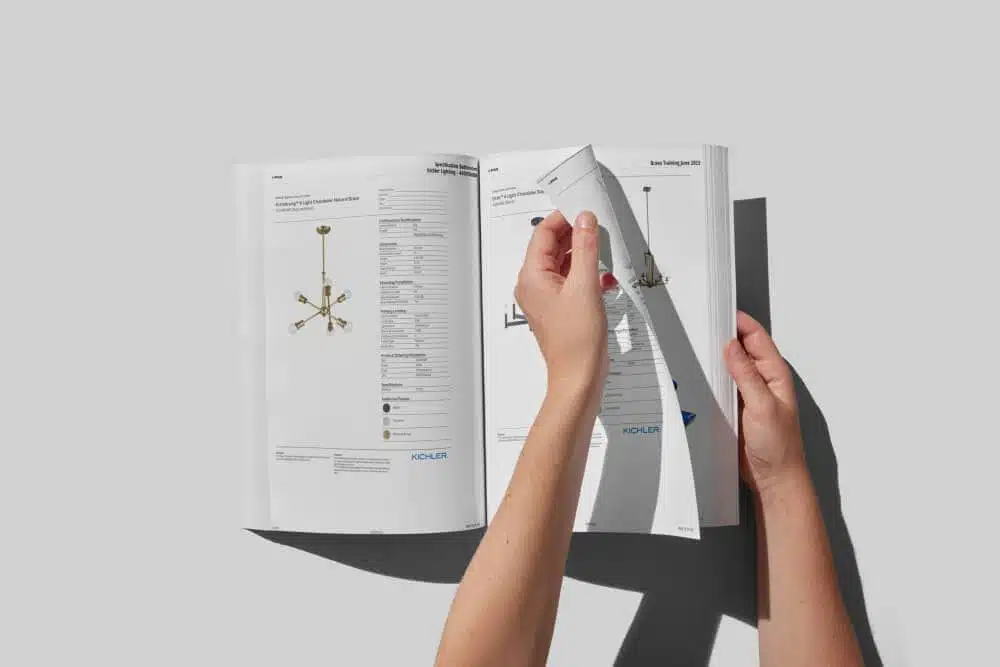 Hands turning the pages of a printed specification book of branded lighting spec sheets, each with product images and a full specification table
