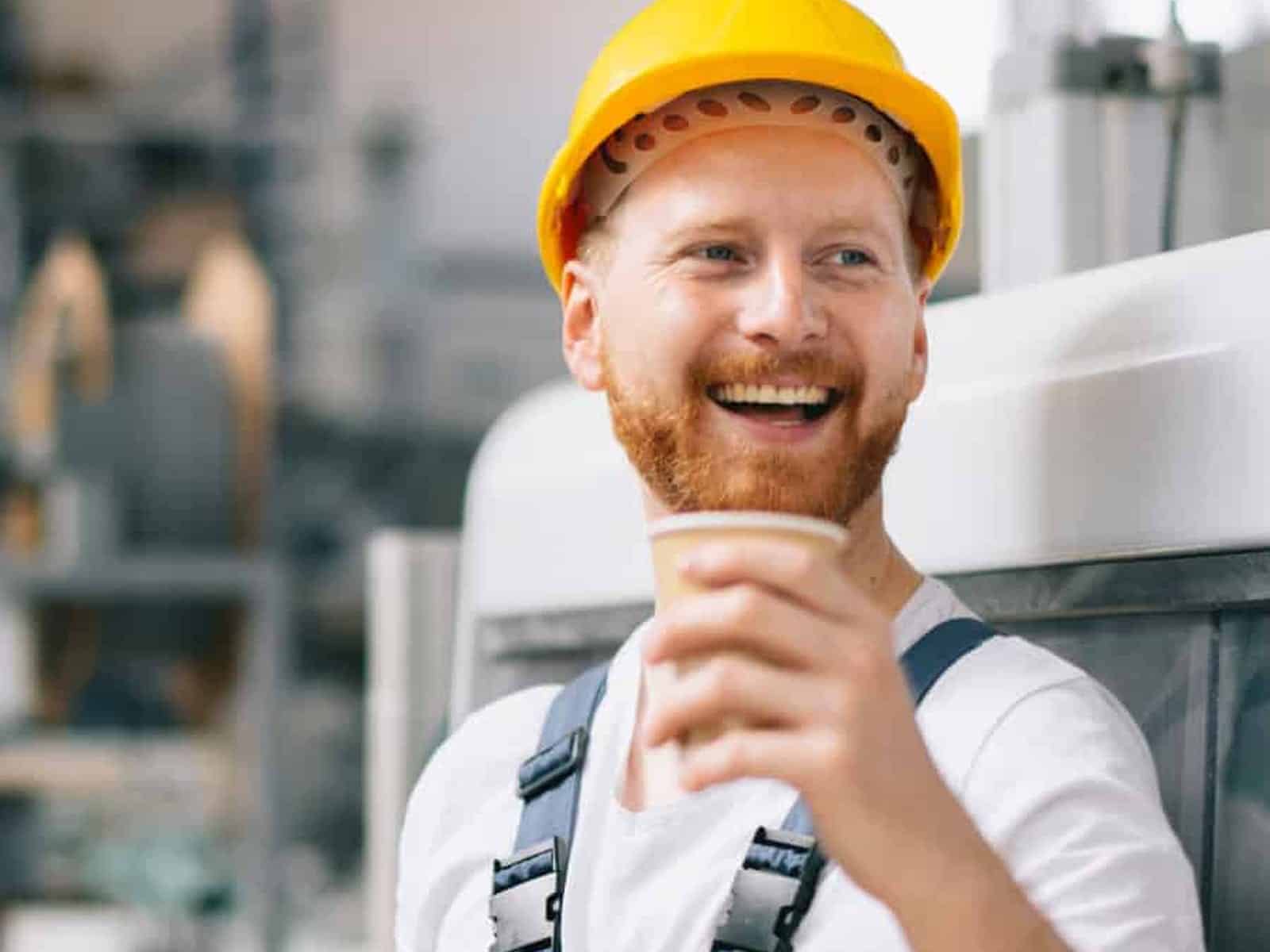 Smiling manufacturing worker in a hard hat holding a coffee on the plant floor